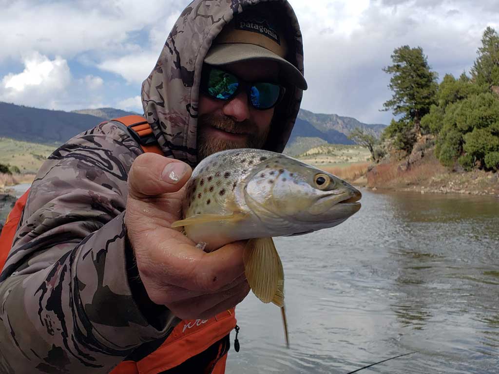 A photo of the mountains in the background and a fly fishing angler squatting in the riverwhile posing with a Trout caught during the fall fishing season