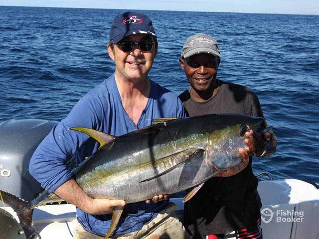 Two men stood on a fishing charter, both wearing sunglasses and baseball caps, while holding a Yellowfin Tuna on a clear day