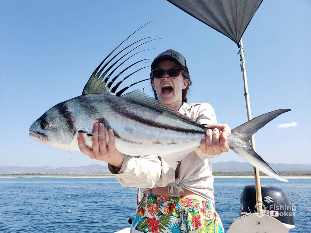 A woman holding a Roosterfish aboard a fishing boat while looking shocked on a clear day with calm waters and blue skies visible behind her