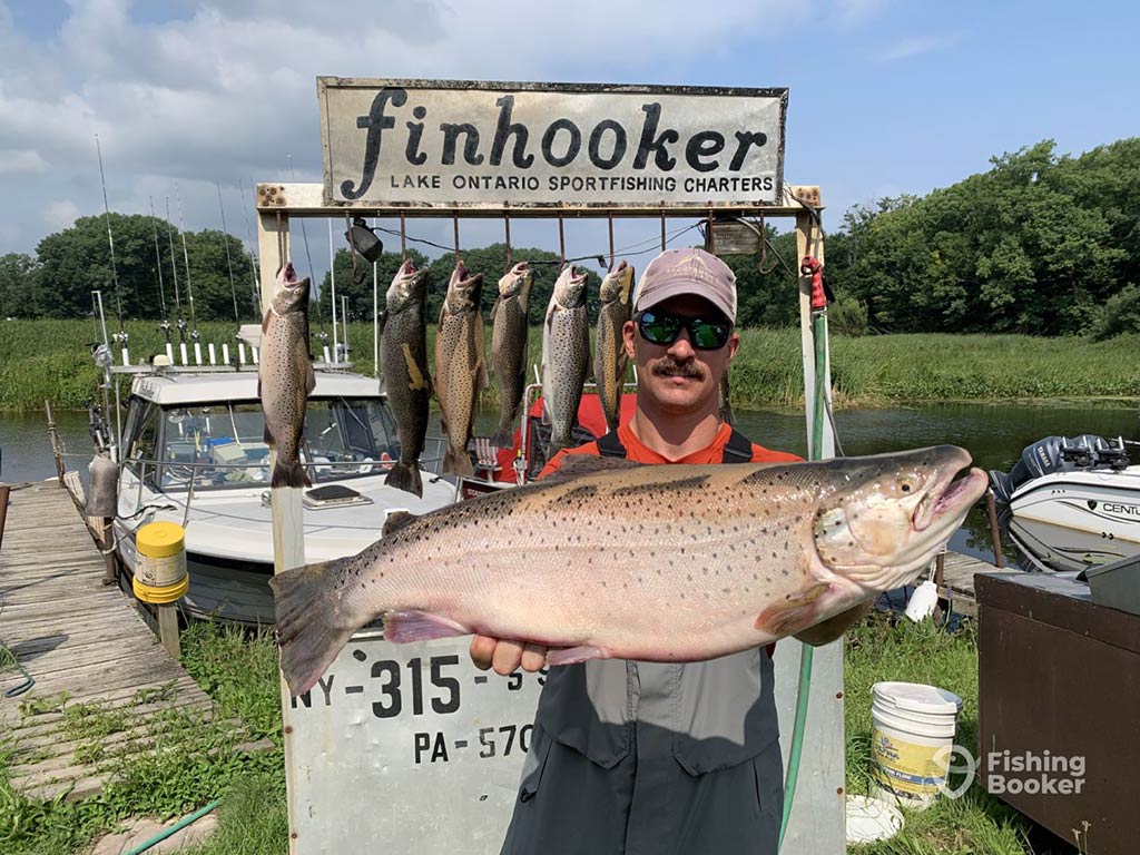 A man standing in front of a wooden sign which says "Finooker Lake Ontario Fishing Charters" and a boat on the water while holding a large Brown Trout with a number of others visible behind him hanging from the sign