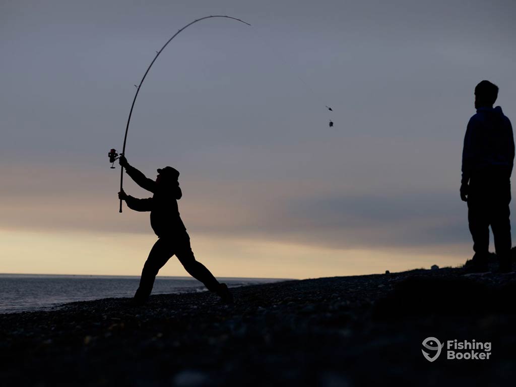 A silhouette of a person casting a heavy-action rod at sunset, with an onlooker stood behind them on a cloudy day