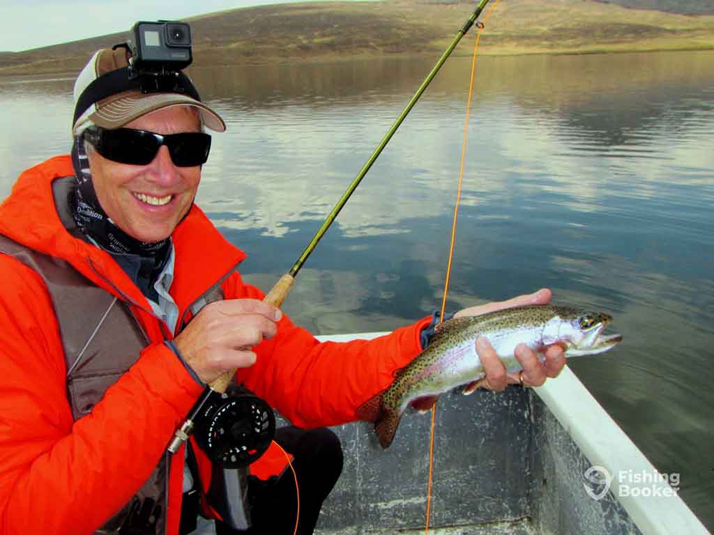 A man in a red jacket, sunglasses, and had with a GoPro camera on it holding a small Trout in one hand and a fly fishing rod in the other aboard a boat on a calm lake