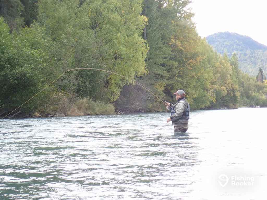 A view across the waster towards a man wading up to his waist in the waters of a river on a cloudy day, as he casts a fly line