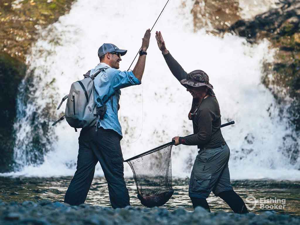 Two men standing in front of a waterfall and high-fiving each other, while holding fly fishing rods and a net with a fish in it