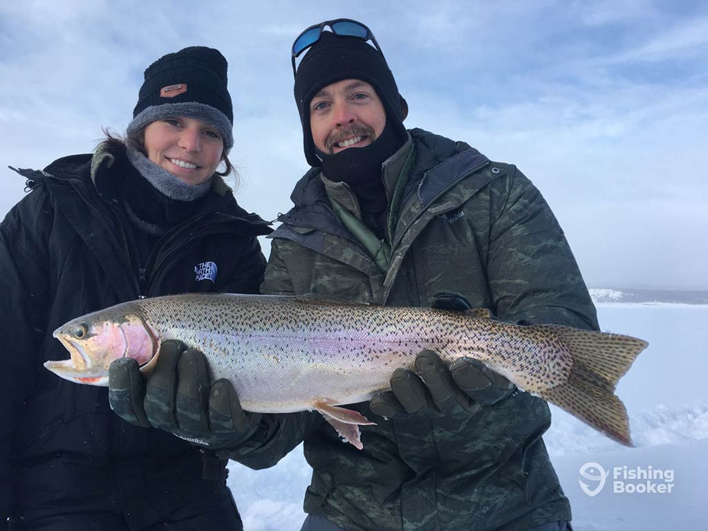 A man and a woman in full winter gear posing with their Cutthroat Trout catch on a frozen lake in Colorado