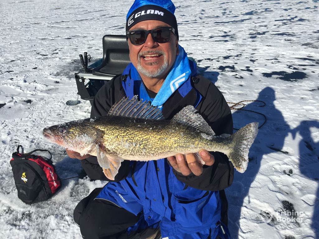 A man in a woolly hat and sunglasses crouches down on a frozen lake in Colorado while holding a Walleye