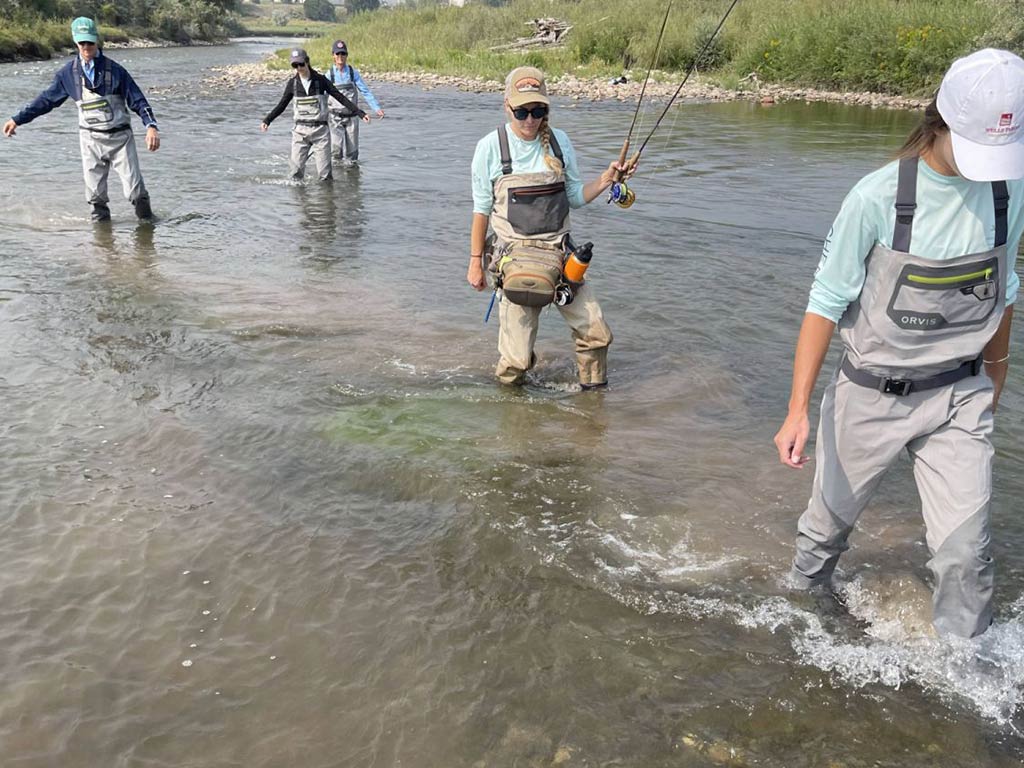 A group of five fly fishing anglers of both genders wading in a row through the shallow waters of the Eagle River in Colorado on a bright day, with the second woman holding a fly fishing rod