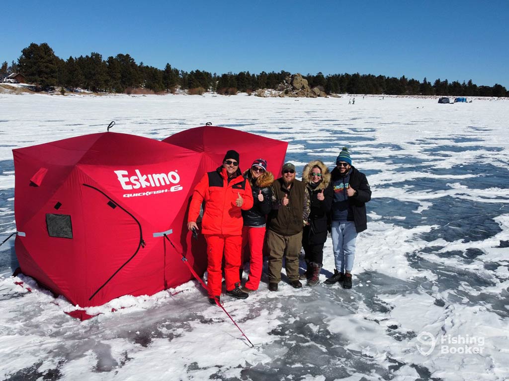 A group of five anglers pose in front of a big, red ice hut on a frozen lake in Colorado on a clear day