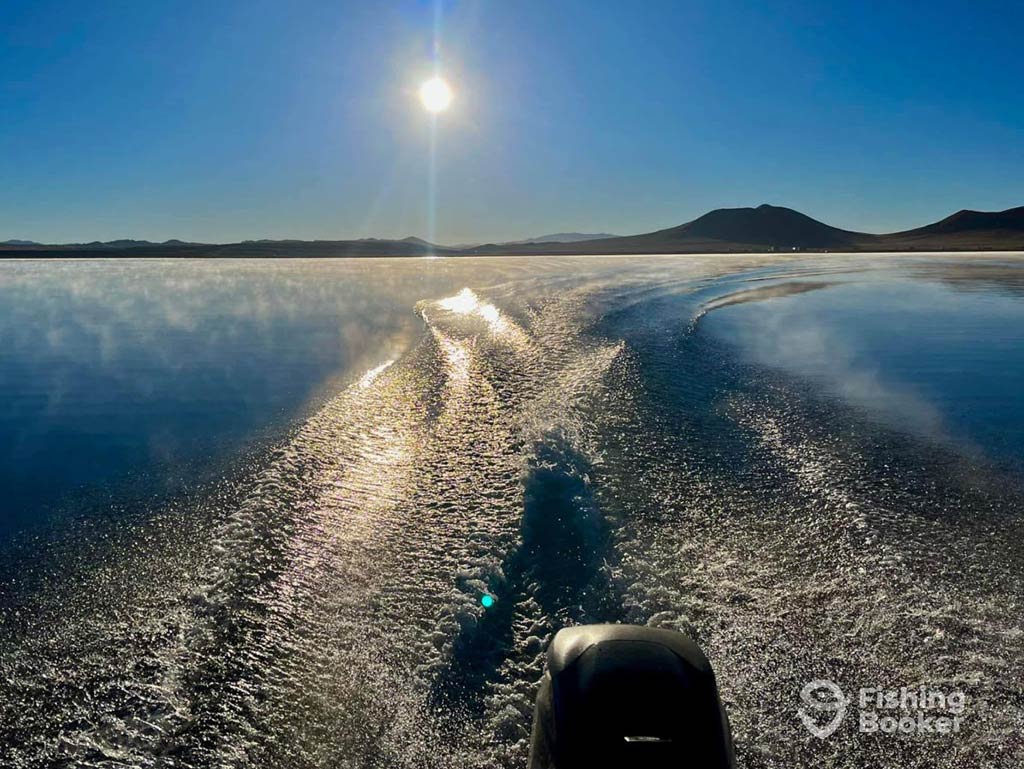 A view out the back of a fishing boat on a lake in Colorado on a clear day, with the sun shining brightly in the distance above some mountains, while the boat's wake is visible behind the engine in the foreground