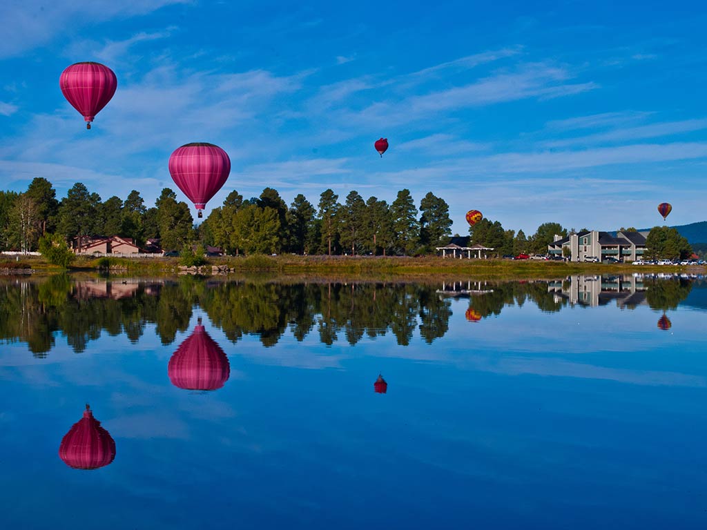 A view across a calm lake in Colorado, reflecting a number of hot air balloons visible above in the clear sky