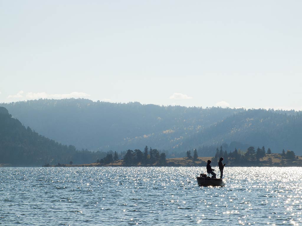 A view across the waters of Eleven Mile Reservoir in Colorado Springs on a clear day, with one boat and two anglers fishing from it in the distance