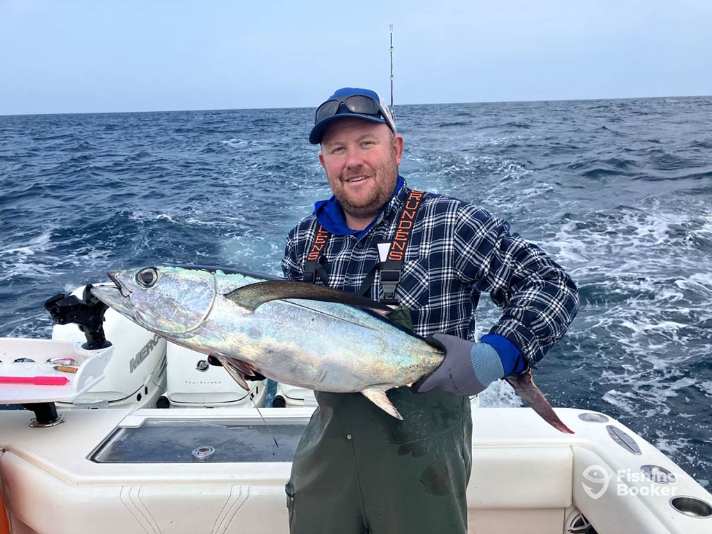 A man in full fishing gear standing on the deck of a boat in the Pacific waters of Washington state while holding a large Albacore Tuna