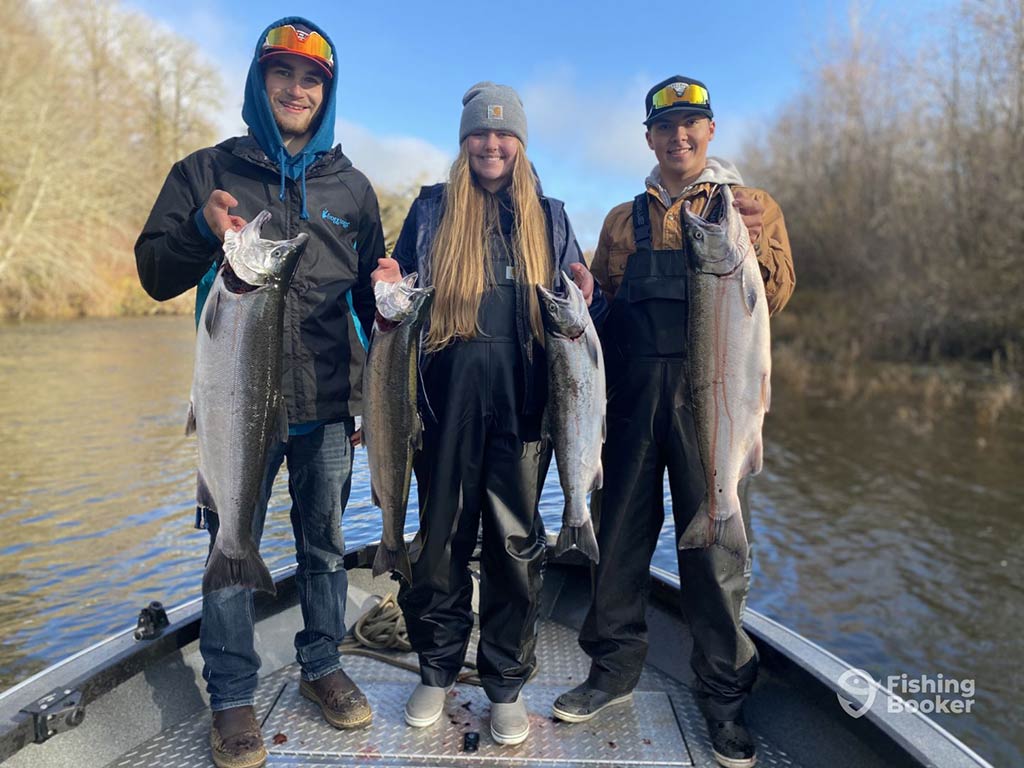 Three youthful anglers, including one woman, holding up four Salmon between them on a river fishing charter in Washington, with fall foliage visible on either side of the river