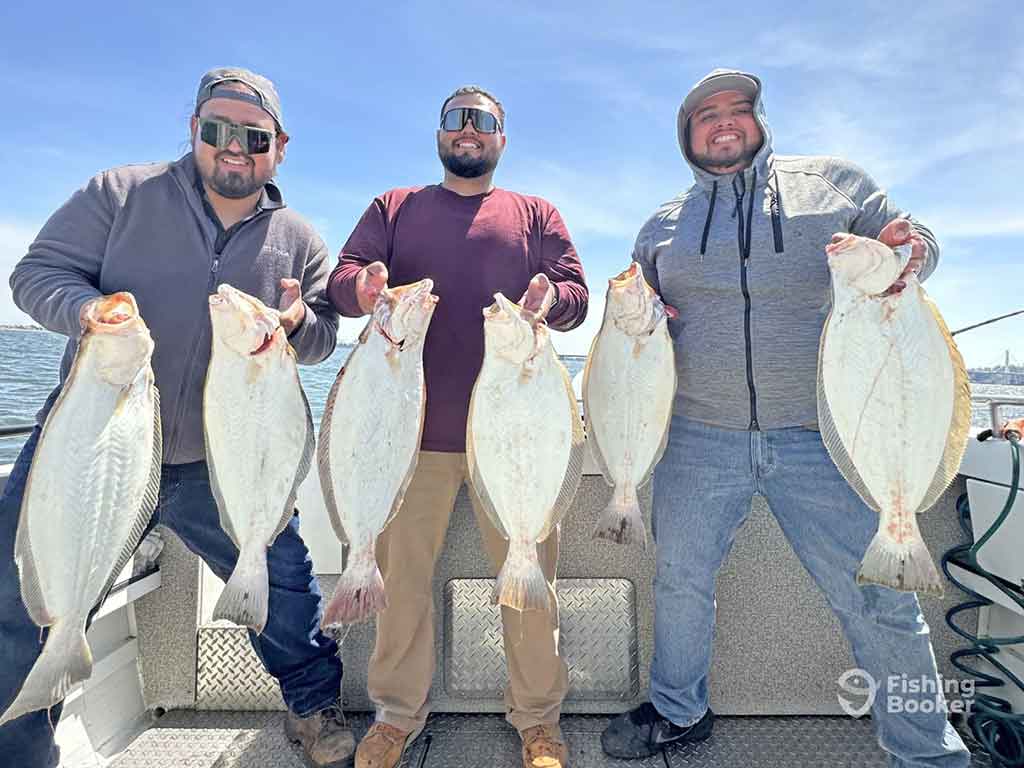Three anglers standing aboard a fishing charter on a sunny day and showing off two Halibuts each