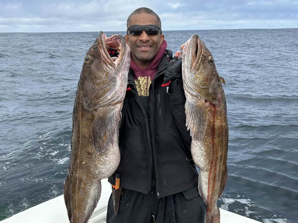 A smiling man in sunglasses standing on a fishing charter and holding two large Lingcods on a cloudy day, with the water visible behind him