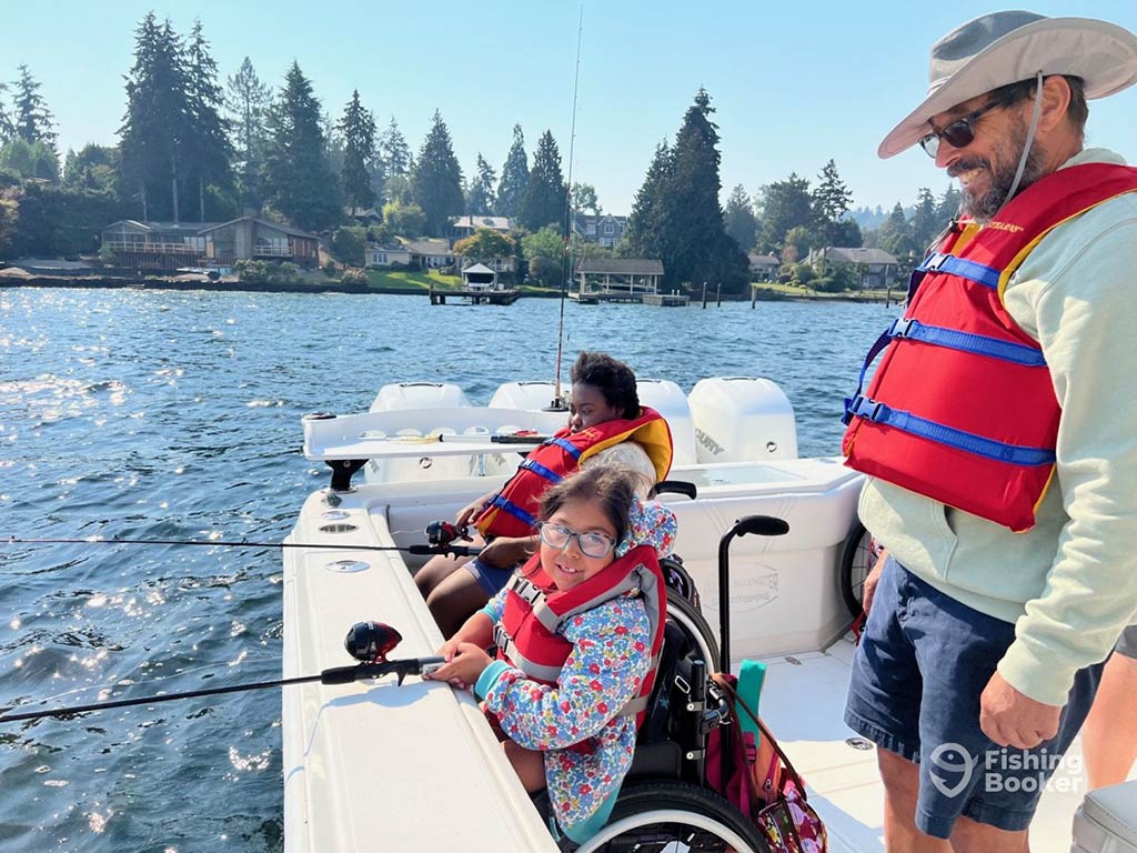 Two young girls in wheelchairs fish over the side of a boat on a suny day in Westport, WA, with a man in a hat standing on the right of the image looking over them