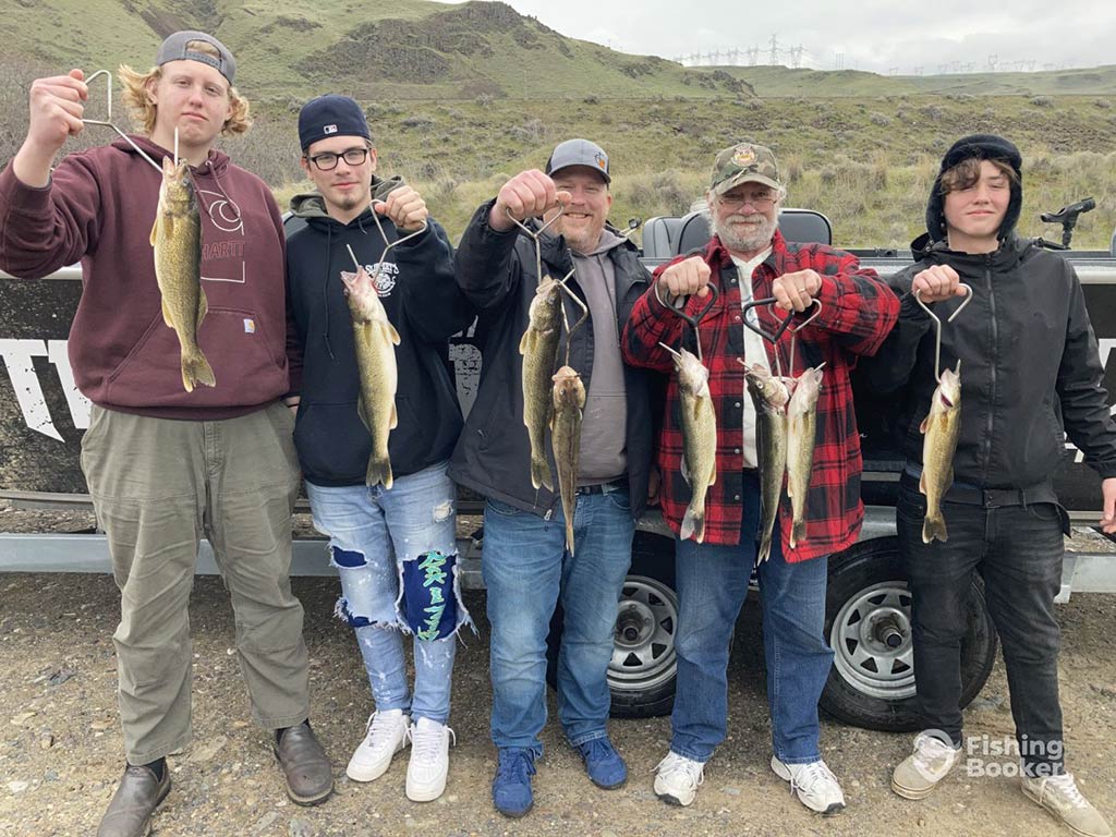 A group of male anglers back on dry land after a successful fishing trip on the Columbia River, showing off a Walleye each, with hills visible behind them on a cloudy day