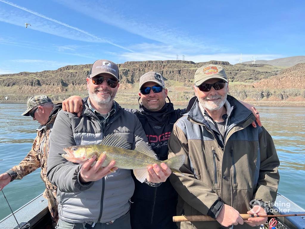 Three men stood on a charter fishing boat on the Columbia River with the one on the left holding a Walleye, with one man standing behind them on a sunny day
