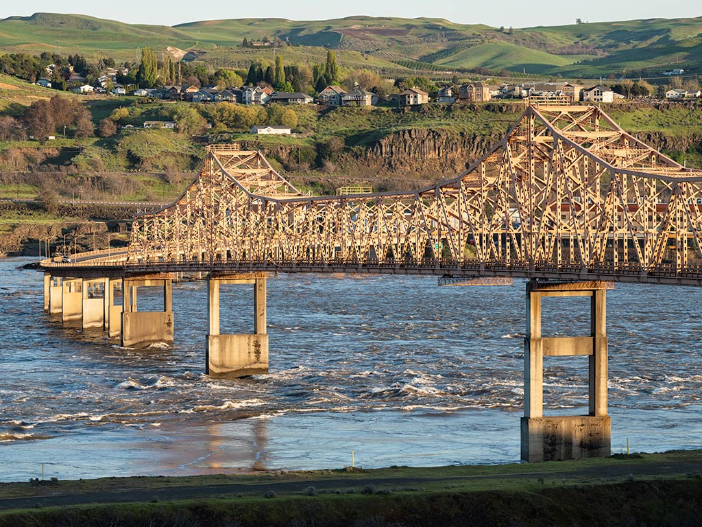 A view of the Dalles Bridge crossing the Columbia River on a clear day, with a green hill dotted with buildings visible in the distance