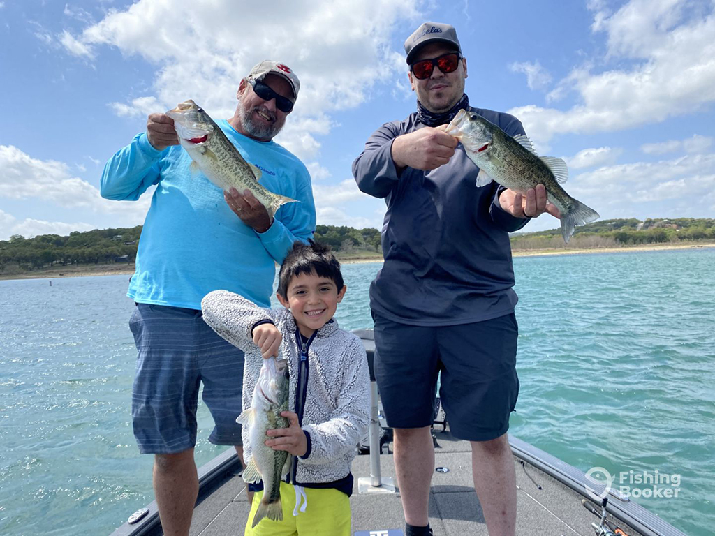 Two adults and a child stand aboard a boat on a lake with the shoreline in the background, holding their catch after Bass fishing in Canyon Lake