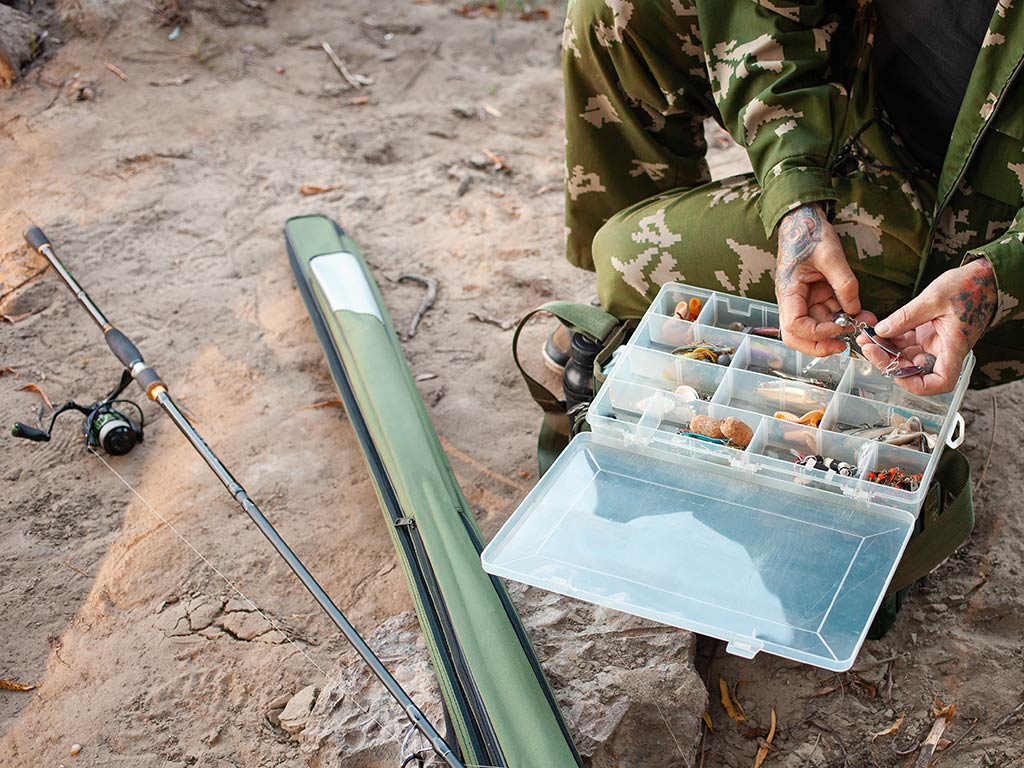 A man crouching down next to a tackle box, while sorting out weights, with a fishing rod and case next to him on the ground