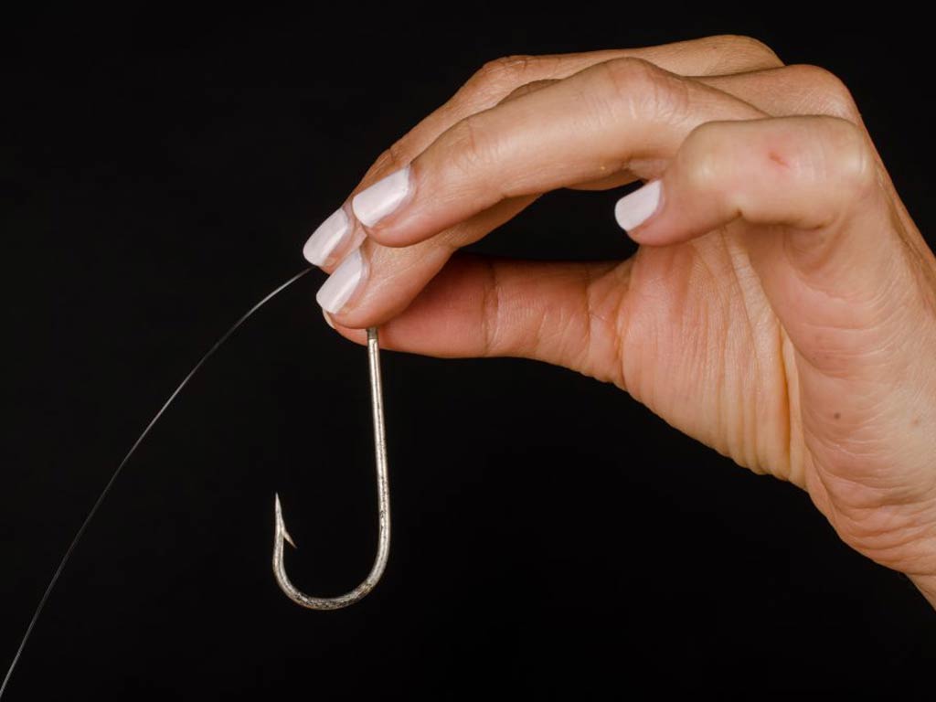 A woman's hand with white nail polish holding an Aberdeen fishing hook against a black background