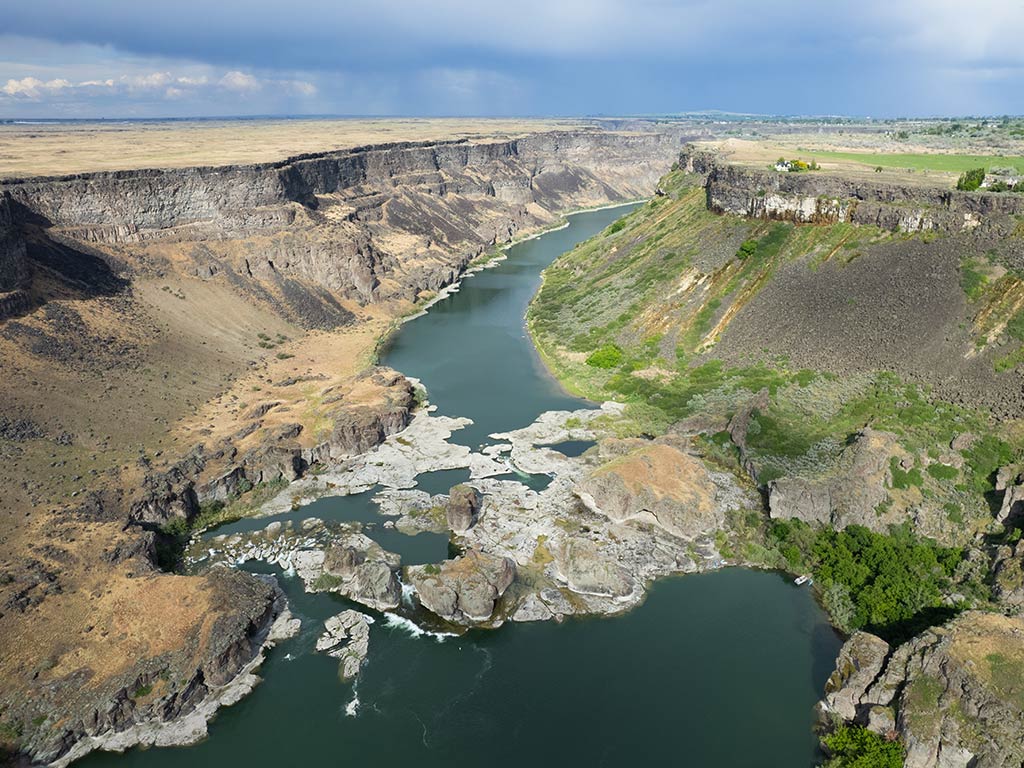 An aerial view of the Snake River Canyon on a cloudy day, with rocks breaking the flow of the river in the middle of the image