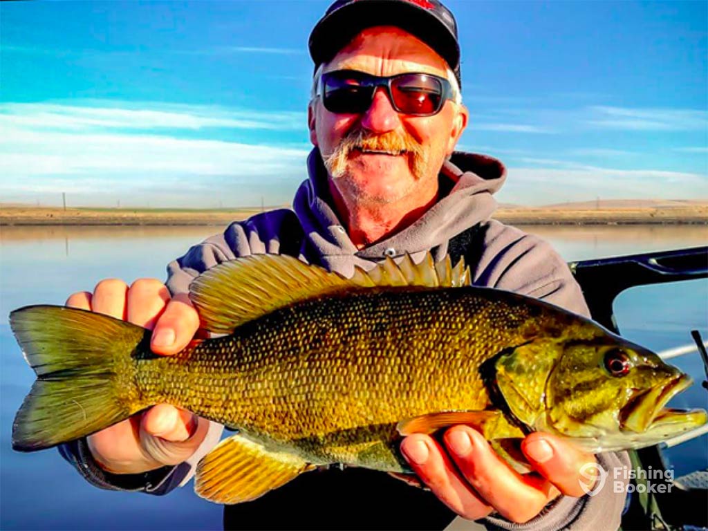 A closeup of a man in sunglasses and a baseball cap smiling as he holds a Smallmouth Bass up to the water on a calm body of water on a clear day