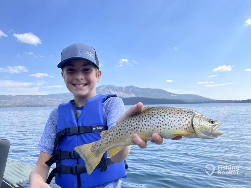 A young boy sitting on a boat, wearing a baseball cap and blue life vest holding a Brown Trout in his left hand wiuth the water visible behind him on a day with sunny intervals