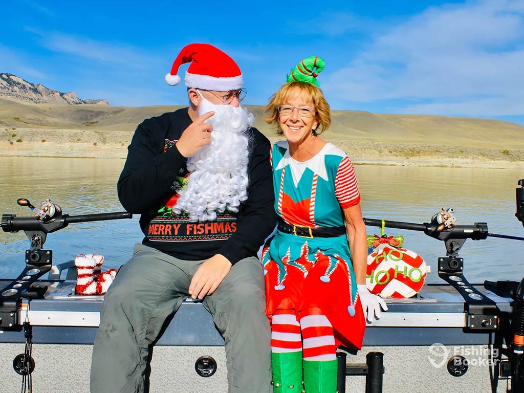 A man and a women dressed in Christmas wear – the man with a Santa hat and beard and the woman dressed as an elf – on a boat on a sunny day, with yellow hills visible across the water behind them