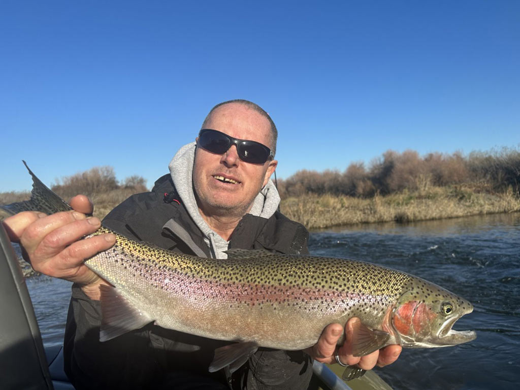 An angler in sunglasses posing with a big Rainbow Trout he caught, the fish's signature pink line and cheeks are clearly visible, while waters and the shoreline can be seen behind the angler.
