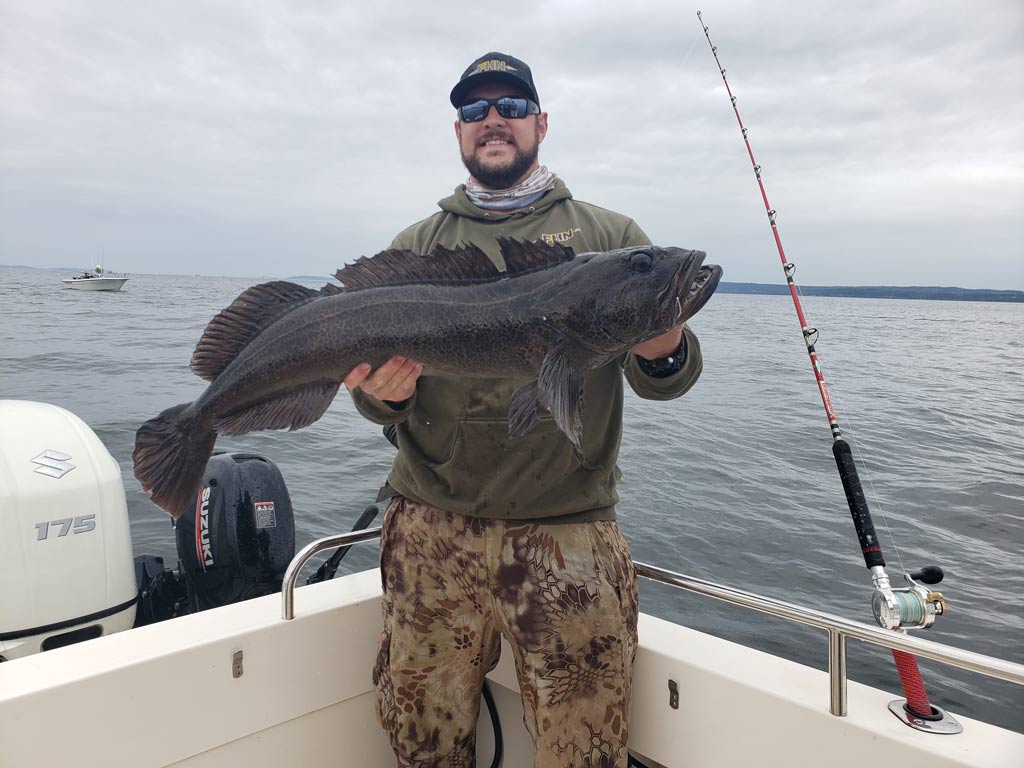 An angler in sunglasses and a hat posing on a charter boat in Washington, holding a big, dark-colored Lingcod he caught, with waters behind him.