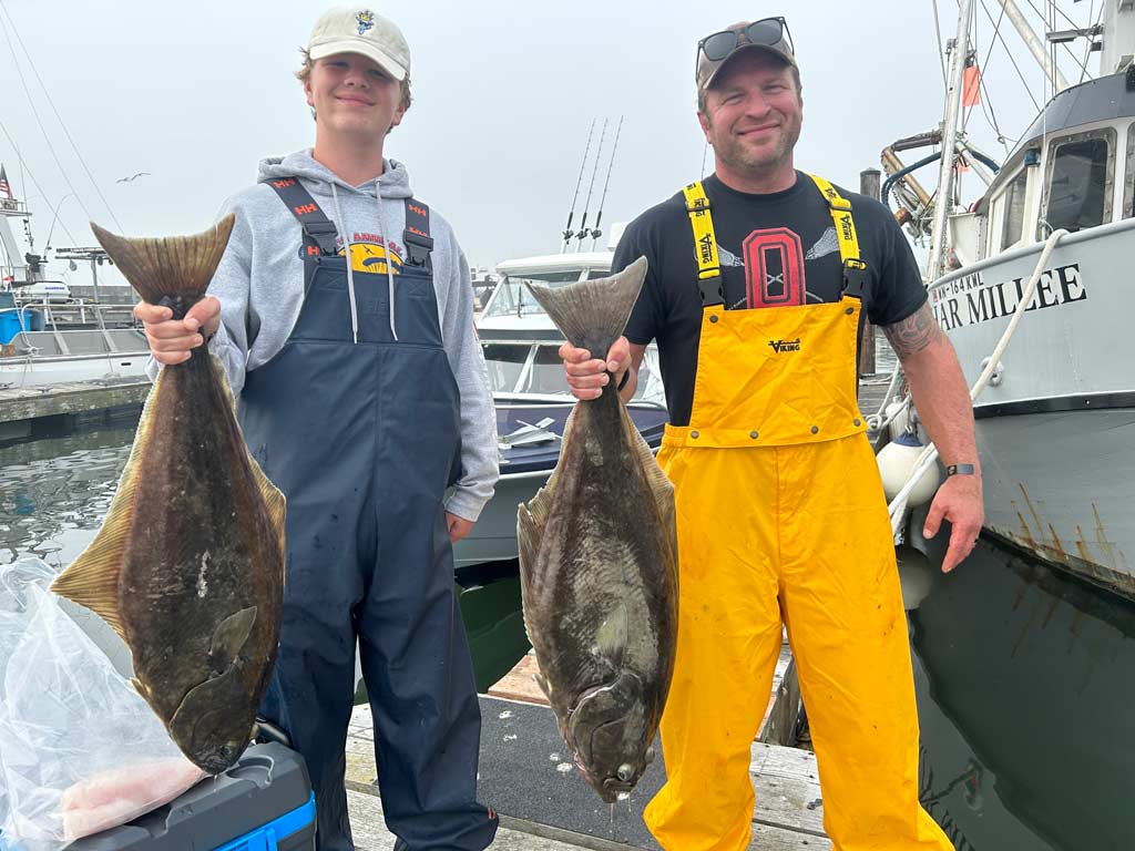 Two anglers in hats and overalls standing on a dock with each holding a Halibut towards the camera, with boats visible behind them.