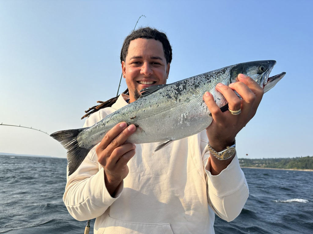 An angler in a white hoodie smiling and holding a Coho Salmon towards the camera with waters and clear skies in the background.