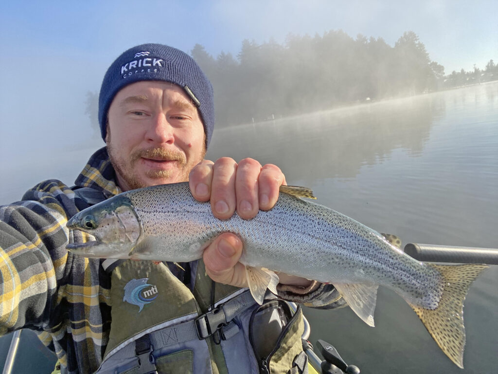 An angler in a winter cap taking a selfie while holding a Rainbow Trout in his left hand towards the camera, with misty waters and trees visible in the background.