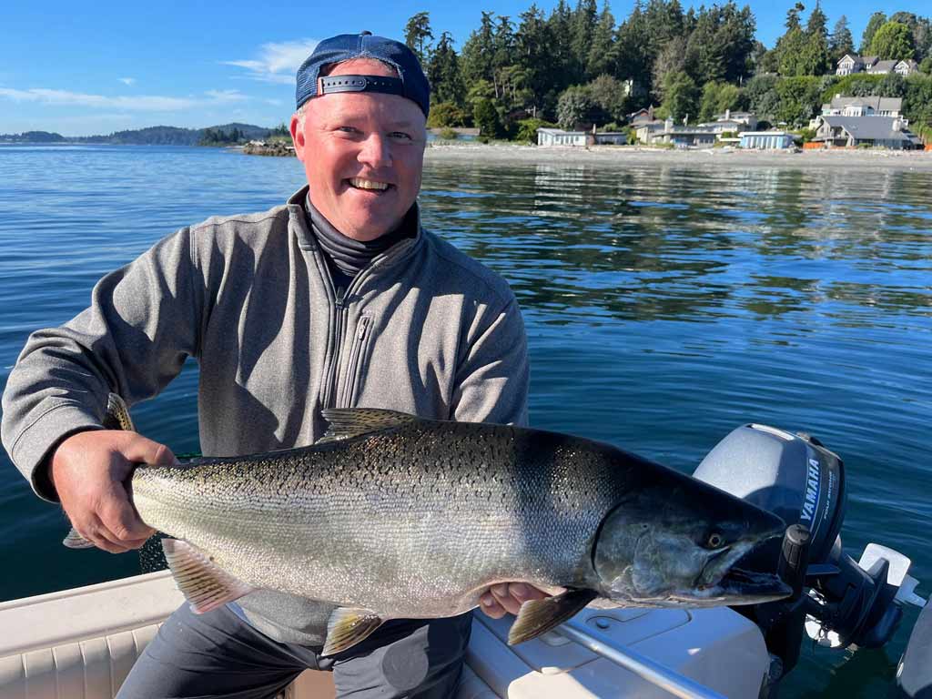 A smiling angler wearing his hat backwards sitting on a boat and holding a massive Chinook Salmon caught fishing during Washington's summer season, with calm waters and shoreline greenery in the background.