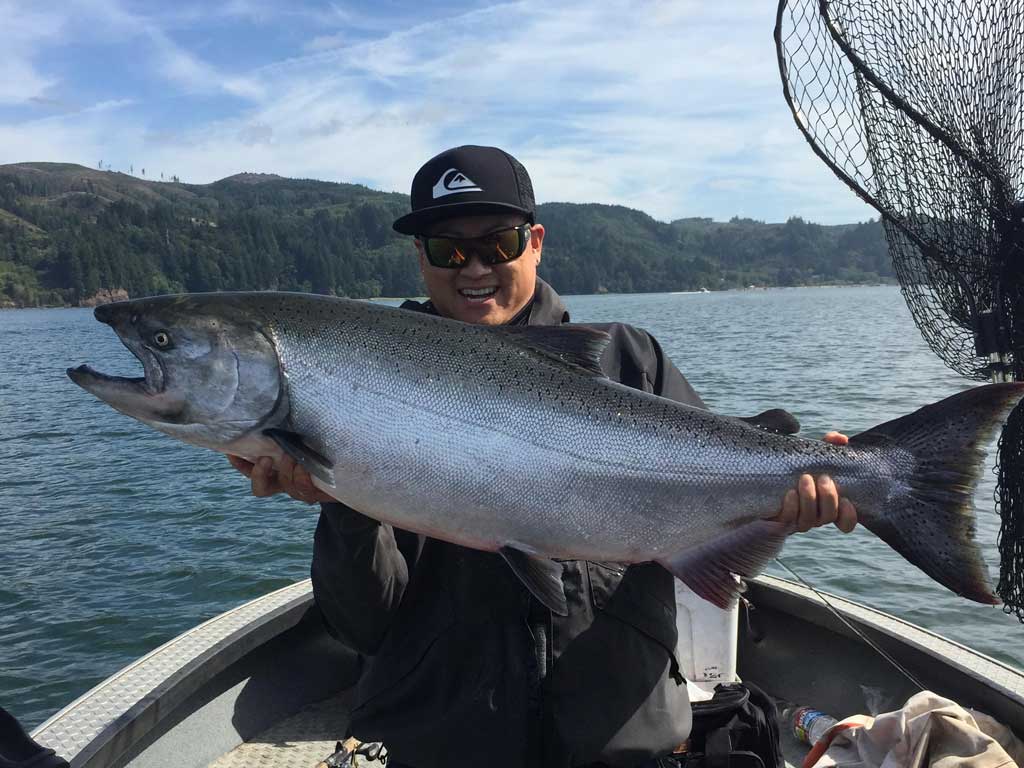 An angler in sunglasses and a hat smiling widely and sitting on a boat while holding a huge King Salmon, with waters and the shoreline visible behind him.