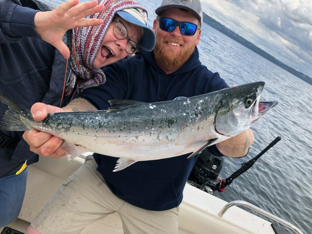 An angler in sunglasses and a hat smiling and holding a Coho Salmon while another person to his left is leaning in for the photo with their mouth open wide.