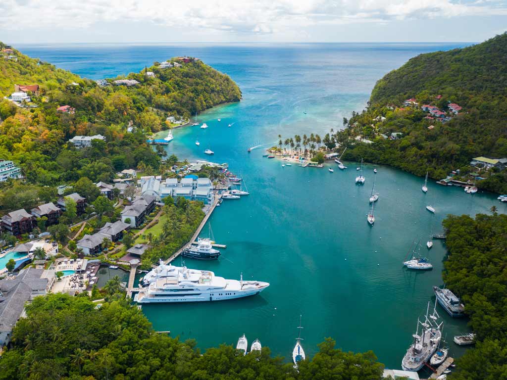 A view of Marigot Bay on Saint Lucia, one of the islands in the Carribean, taken from above the bay with docked ships visible on azure waters that stretch towards the horizon.
