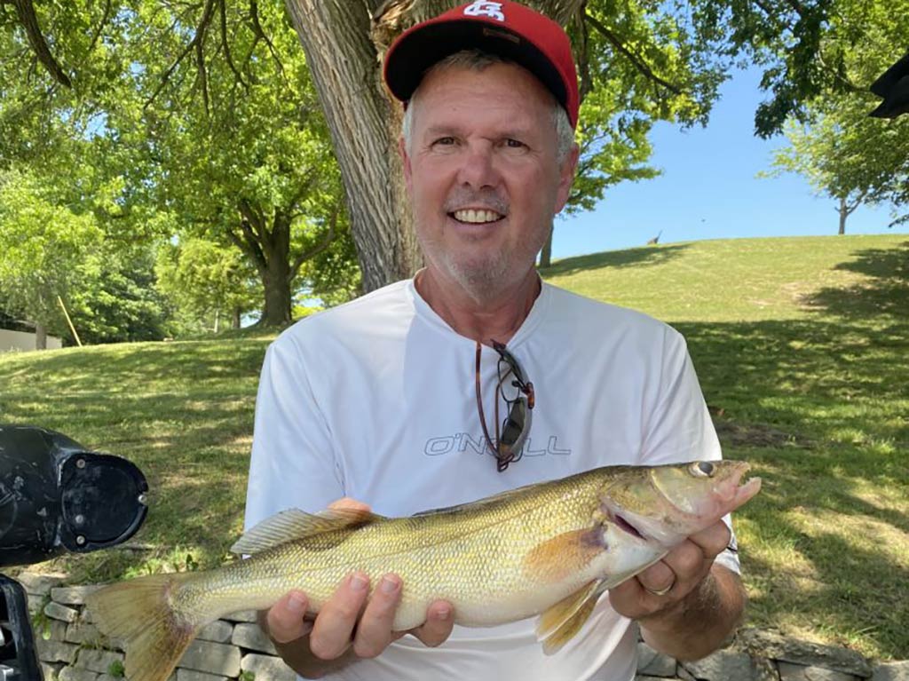 A man in a red baseball cap smiles as he holds a Walleye back on shore and in the shade of some green trees on a bright day