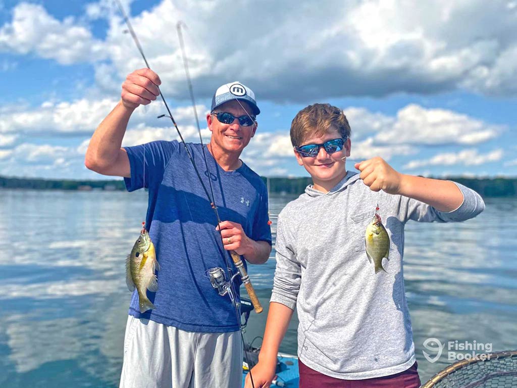 A man and teenage boy pose with two small Bluegills, which they're holding by their fishing lines aboard a fishing charter on a day with sunny intervals