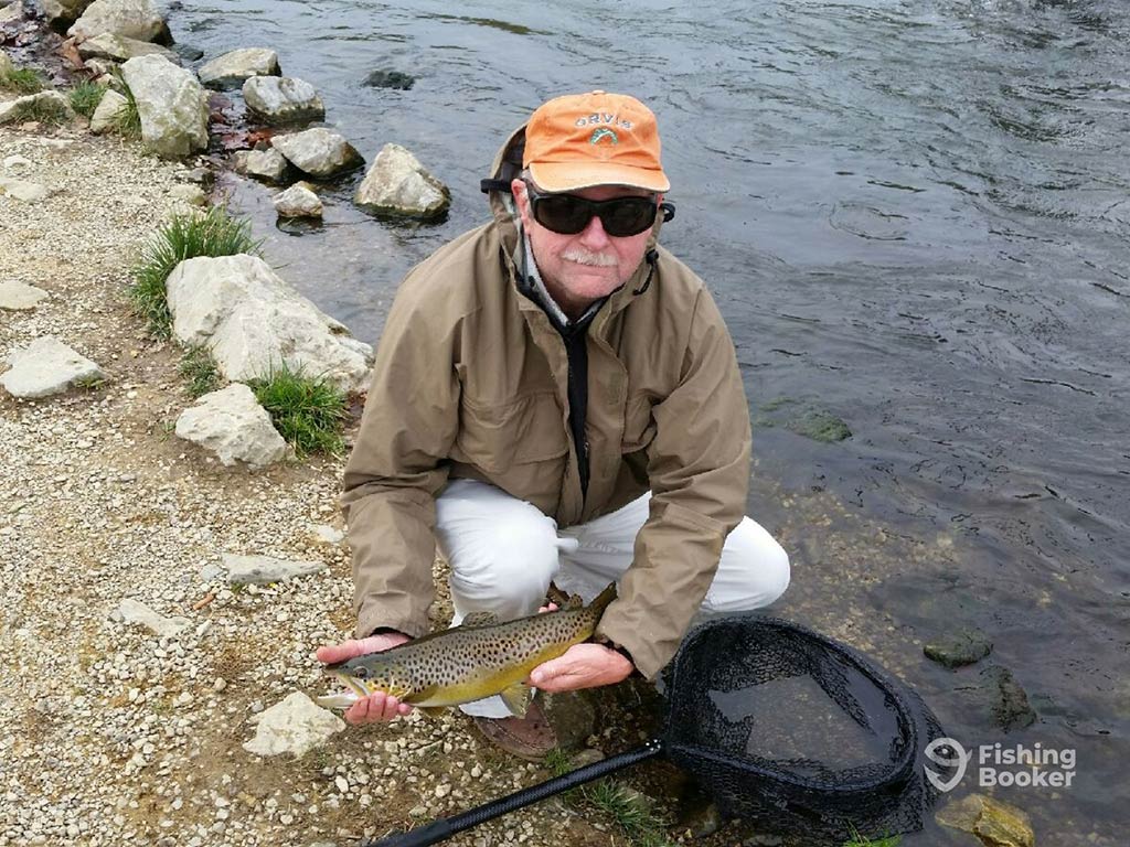 An elderly angler in an orange baseball cap and sunglasses, crouching down next to a river while holding his Brown Trout catch