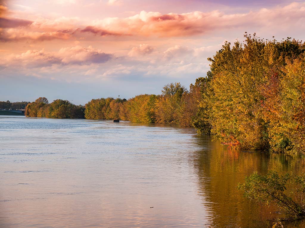 A view along a shoreline of the White River in Indiana at sunset, with fall colors dominating among the trees on the right of the image
