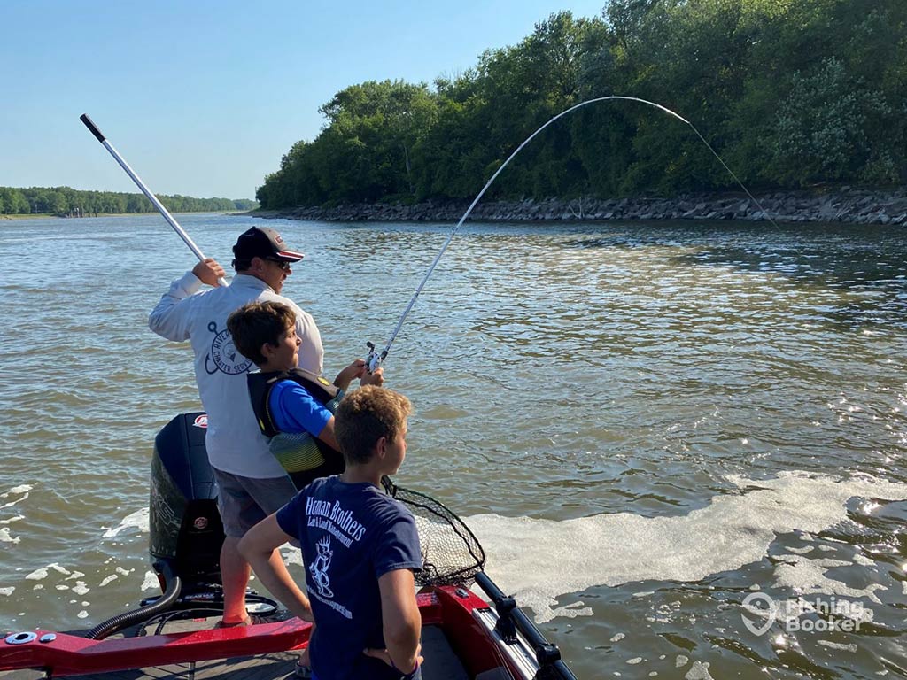 A child struggles with a bent rod over the side of a fishing charter on a river in Indiana, as a man assists behind him with a net, while another child looks on in the foreground on a clear day