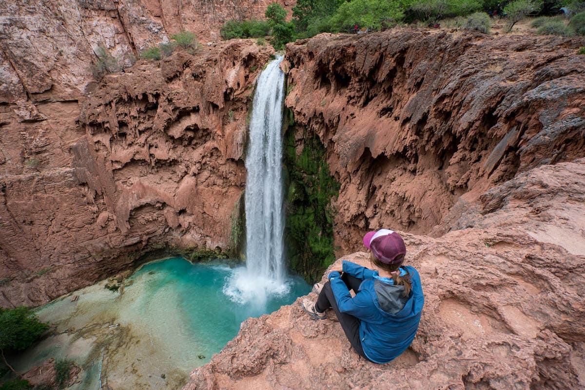 Woman sitting on rock ledge looking down over Mooney Falls in Havasupai with turquoise water and red rock cliffs