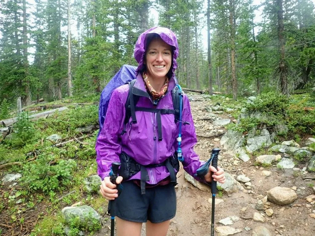 Woman wears purple Arcteryx rain jacket while hiking on rocky trail in the rain