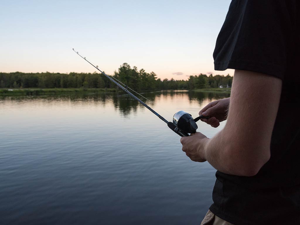 A view from behind of a man's arm holding a fishing rod set up for spin fishing at dusk on a clear day, with calm waters leading to a shoreline visible in the distance