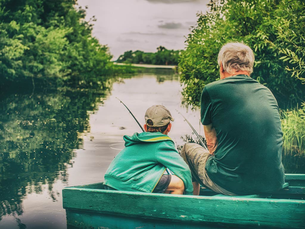 A view from behind of a grandfather and grandson sitting in a small boat on a river surrounded by greenery, with each casting into the water on a cloudy day