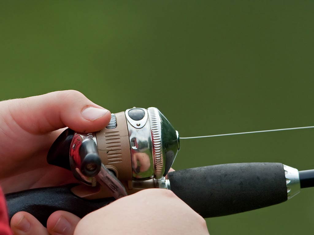 A closeup of a hand holding a small spinning reel on a fishing rod against a green background