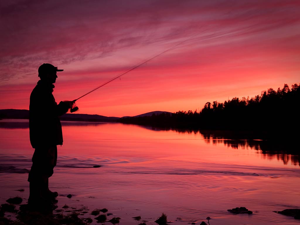 A slhouette of a man fishing with a spinning rod and reel combo on a lake against the bright red light of the sun setting